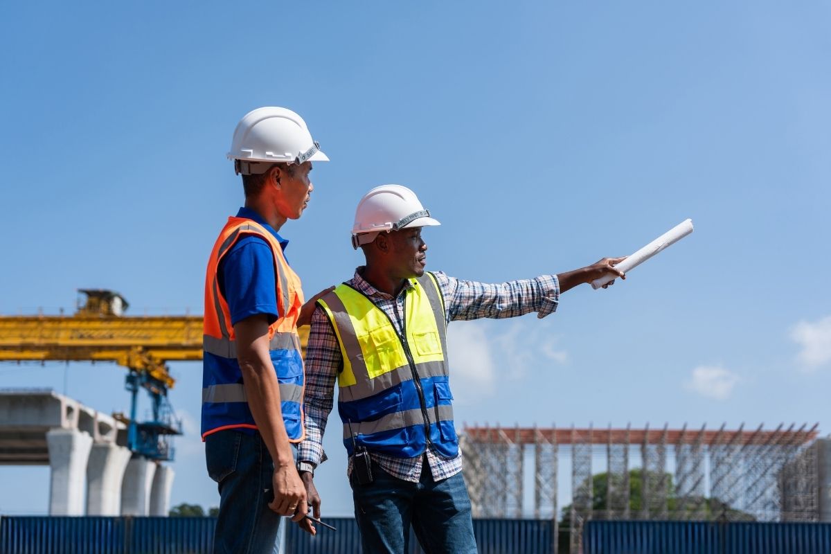 Two licensed contractors in hard hats and safety vests reviewing blueprints on a construction job site