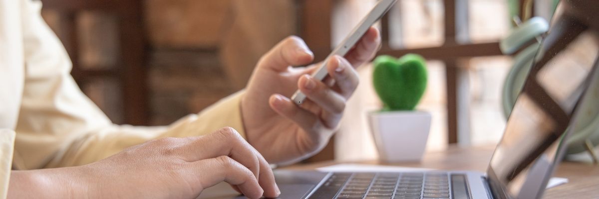 Person using a smartphone while typing on a laptop to complete an online application form